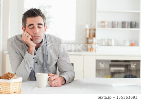 Man in early 30s resting head on hand, sipping from white mug at bright kitchen table 135363693