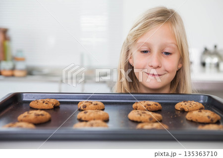 Child girl leaning, gazing at baking tray of chocolate chip cookies on kitchen counter, copy space 135363710