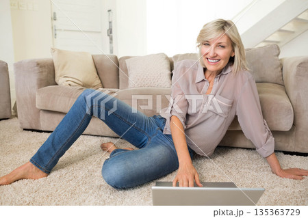 Mature adult woman sitting on carpet in living room using silver laptop, leaning against beige sofa Mature adult woman sitting on carpet in living room using silver laptop, leaning against beige sofa 135363729