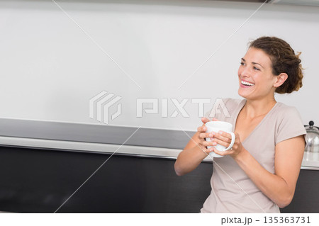 Woman leaning against dark countertop in kitchen while holding white ceramic mug, copy space Woman leaning against dark countertop in kitchen while holding white ceramic mug, copy space 135363731