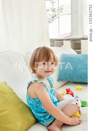 Toddler girl sitting cross-legged on white sofa in living room amid colorful alphabet blocks 135363752