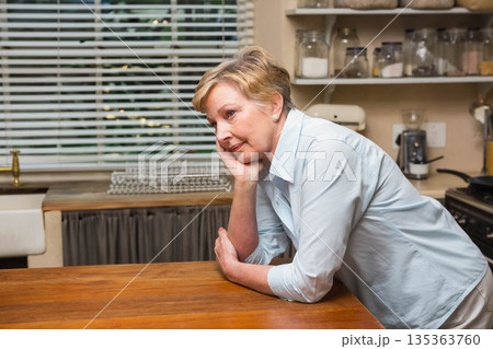 Senior woman leaning on countertop by sink in kitchen, resting head on hand, gazing ahead 135363760