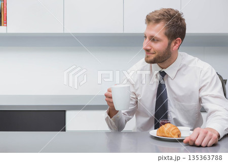 Man holding ceramic mug while sitting at office kitchenette counter with croissant, copy space Man holding ceramic mug while sitting at office kitchenette counter with croissant, copy space 135363788