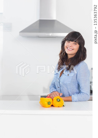 Woman leaning on kitchen island smiling with yellow peppers under range hood, copy space 135363792