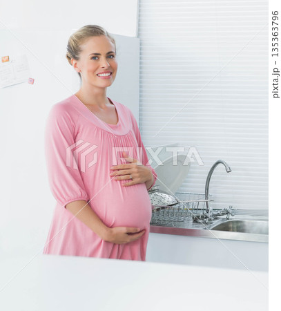 Mid-adult pregnant woman in maternity dress standing near sink at home with dish rack and magnets 135363796