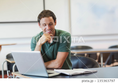 Male student studying at wooden desk in classroom using silver laptop, notebook and pen 135363797