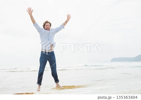 Man wearing blue button-down shirt and cuffed jeans jumping in shallow ocean water at shoreline 135363804