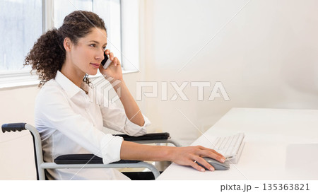 Female worker speaking on smartphone while clicking mouse on office desk in wheelchair, copy space 135363821