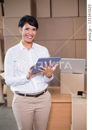 Woman smiling and tapping tablet while standing at shipping station with table, laptop and boxes 135363829