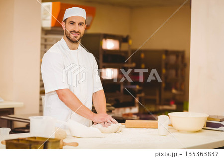 Male baker wearing white uniform kneading dough on floured countertop in bakery with rolling pin Male baker wearing white uniform kneading dough on floured countertop in bakery with rolling pin 135363837