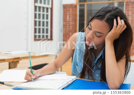Asian woman leaning on left hand and writing in binder with green pen in study area 135363900