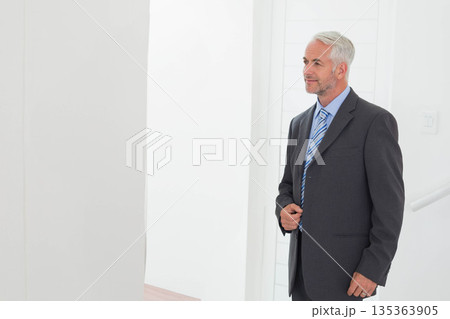 Senior male professional buttoning grey suit jacket in office hallway near paneled wall, copy space Senior male professional buttoning grey suit jacket in office hallway near paneled wall, copy space 135363905
