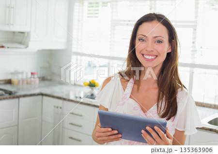 Woman wearing floral apron holding tablet checking recipe in kitchen at countertop with spice jars Woman wearing floral apron holding tablet checking recipe in kitchen at countertop with spice jars 135363906