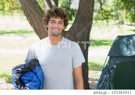 Man standing under tree next to pitched tent at campground holding blue and black sleeping bag Man standing under tree next to pitched tent at campground holding blue and black sleeping bag 135363911