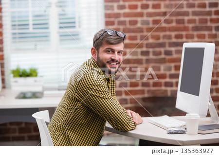 Man wearing eyeglasses smiling while leaning at desk in office with computer coffee cup brick wall 135363927