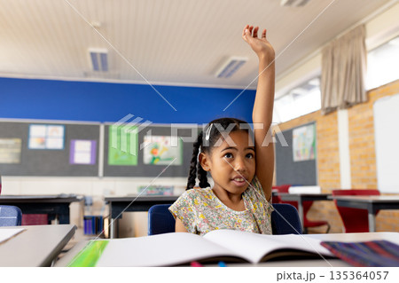 Young African American girl raising hand at desk in elementary school classroom with open workbook Young African American girl raising hand at desk in elementary school classroom with open workbook 135364057