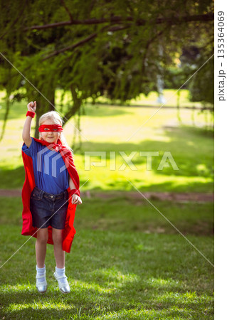 Girl is standing on sunlit park lawn under tree, wearing red superhero cape and mask Girl is standing on sunlit park lawn under tree, wearing red superhero cape and mask 135364069