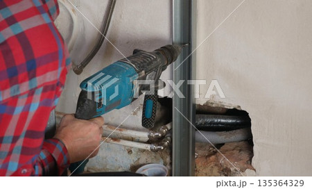 A worker in a checkered shirt is drilling a hole in a metal profile installed in the wall structure with a large drill during renovation work in the building. 135364329