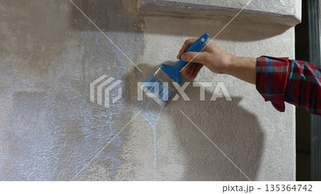 A painter's hand holds a blue brush and applies liquid primer to a textured plastered wall, priming a wall in a room with uneven texture using a paintbrush A painter's hand holds a blue brush and applies liquid primer to a textured plastered wall, priming a wall in a room with uneven texture using a paintbrush 135364742