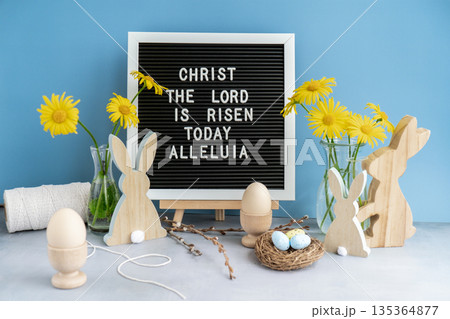 Phrase CHRIST THE LORD IS RISEN TODAY ALLELUIA traditional greeting to christian holiday. Festive mood composition with bunnies and yellow flowers on pastel blue background. Sustainable natural Phrase CHRIST THE LORD IS RISEN TODAY ALLELUIA traditional greeting to christian holiday. Festive mood composition with bunnies and yellow flowers on pastel blue background. Sustainable natural 135364877