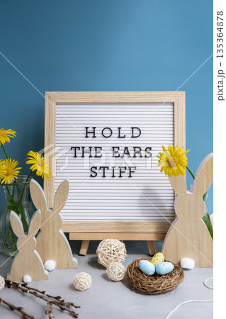 Letter board with message HOLD THE EARS STIFF on festive blue background and eco-friendly zero waste natural decoration. Yellow daisies flowers. Bunny egg. Holiday greeting Letter board with message HOLD THE EARS STIFF on festive blue background and eco-friendly zero waste natural decoration. Yellow daisies flowers. Bunny egg. Holiday greeting 135364878