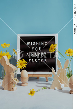 Letter board with message WISHING YOU A HAPPY EASTER on festive blue background and eco-friendly zero waste natural decoration. Yellow daisies flowers. Bunny egg. Holiday greeting 135364883