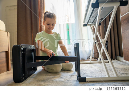 Young girl adjusting chair to piano in home interior before music practice. Preparation for lesson, independence, responsibility, and everyday childhood learning routine. 135365512