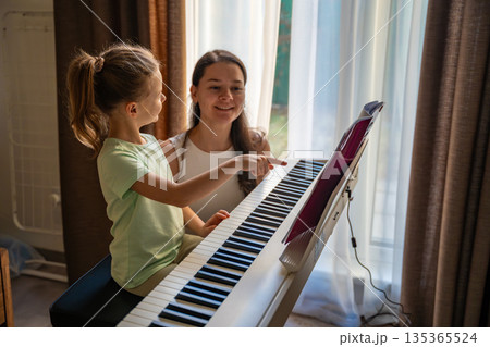 Young girl learning piano with teacher during private music lesson at home. Individual piano education, child development, and focused learning process. Young girl learning piano with teacher during private music lesson at home. Individual piano education, child development, and focused learning process. 135365524