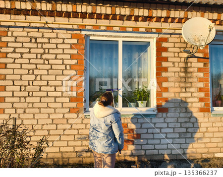 A woman stands outside and cleans a house window with a squeegee on a sunny day. A rear view of a woman cleaning a house window while standing outside. Ordinary household chores. A woman stands outside and cleans a house window with a squeegee on a sunny day. A rear view of a woman cleaning a house window while standing outside. Ordinary household chores. 135366237