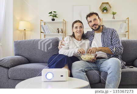 Smiling couple on sofa watching movie with projector at home, eating popcorn 135367273