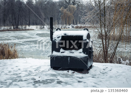 A large black metal stove stands on snow near a frozen pond in winter. 135368460