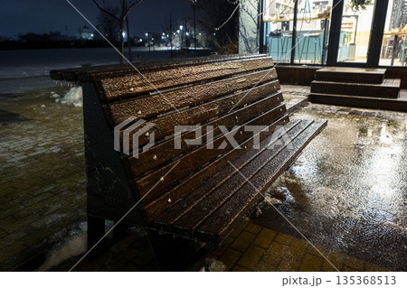 Icy wooden bench in a winter park at evening. Frozen texture and snow in the dark. Icy wooden bench in a winter park at evening. Frozen texture and snow in the dark. 135368513