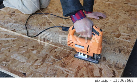 A partially visible white man kneeling with an orange electric jigsaw in his right hand and sawing a sheet of OSB plywood lying on the ground, splitting an OSB board into two pieces using a jigsaw A partially visible white man kneeling with an orange electric jigsaw in his right hand and sawing a sheet of OSB plywood lying on the ground, splitting an OSB board into two pieces using a jigsaw 135368669