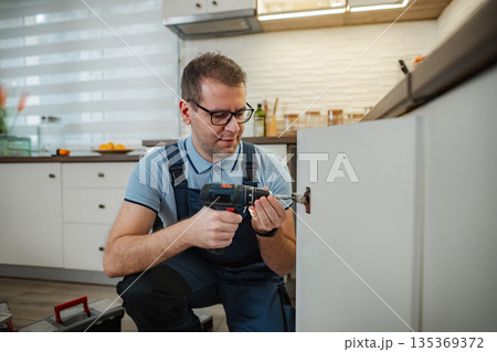 Handyman repairing kitchen cabinet hinge with power drill 135369372