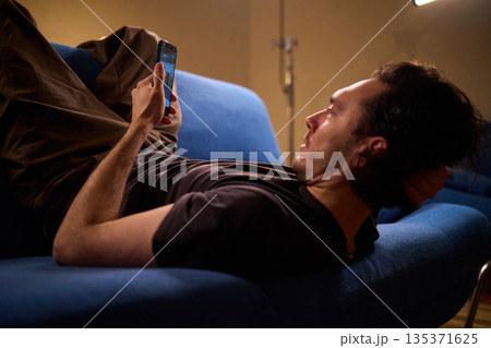 Chilled man on sofa, Casual individual reclining on sofa engrossed with mobile device, Unwinding man sits comfortably on sofa focused on his smartphone amid cozy modern decor 135371625