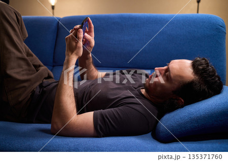 Chilled man on sofa, Casual individual reclining on sofa engrossed with mobile device, Unwinding man sits comfortably on sofa focused on his smartphone amid cozy modern decor 135371760