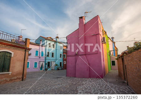 Colorful houses of Burano island near Venice showcasing vibrant pastel facades, narrow streets, and traditional Italian architecture in a quiet residential square under soft daylight sky 135372260