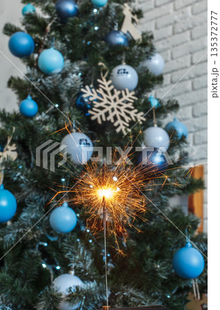 Sparkler shines in front of decorated Christmas tree with blue ornaments in indoor setting Sparkler shines in front of decorated Christmas tree with blue ornaments in indoor setting 135372777