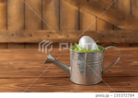 Silver egg in a watering can with green grass placed on a wooden table in a rustic setting 135372784