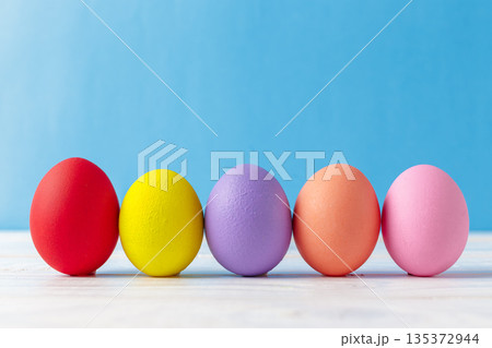 Colorful eggs arranged in a line on a light surface against a blue background Colorful eggs arranged in a line on a light surface against a blue background 135372944