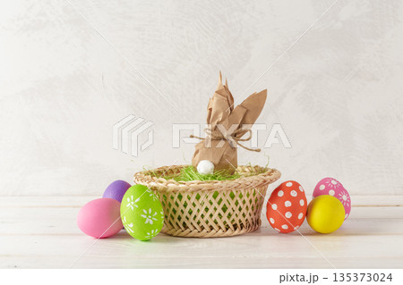 Colorful eggs and bunny wrapped in paper sit in a basket on a bright table surface 135373024