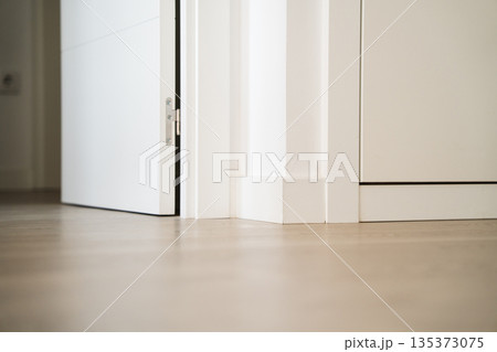 Low angle view of white interior door opening onto light wooden floor, minimalist apartment detail with baseboards and clean modern design Low angle view of white interior door opening onto light wooden floor, minimalist apartment detail with baseboards and clean modern design 135373075