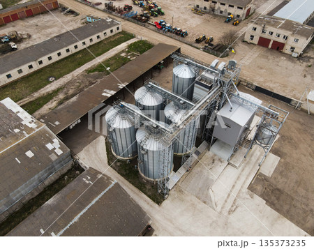 Aerial drone shot of industrial farm grain facility with metal storage silos, conveyor system and warehouse roofs, rural logistics 135373235