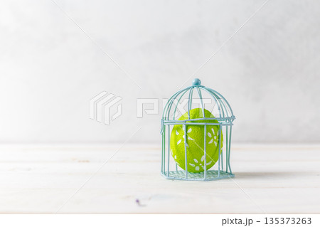 Colorful egg inside a wire cage on a wooden surface in a bright space Colorful egg inside a wire cage on a wooden surface in a bright space 135373263