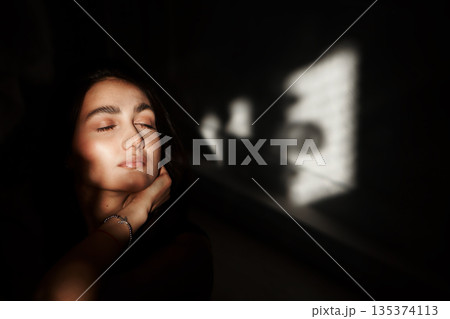 A woman with black hair wakes up in the morning in a white blanket and catches the rays of the sun. The portrait is creative, with brown eyes. High quality photo 135374113