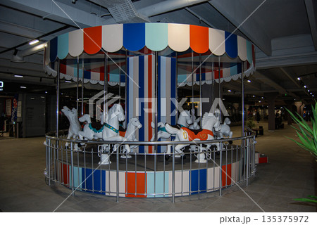 Colorful indoor carousel with white horses. Indoor carousel featuring white horses with colorful saddles, set in a modern, spacious environment. Colorful indoor carousel with white horses. Indoor carousel featuring white horses with colorful saddles, set in a modern, spacious environment. 135375972