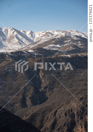 Snow-covered mountain ridges, deep gorge landscape in Armenia with Tatev Monastery visible in the distance, clear winter sky, dramatic natural relief Snow-covered mountain ridges, deep gorge landscape in Armenia with Tatev Monastery visible in the distance, clear winter sky, dramatic natural relief 135376821