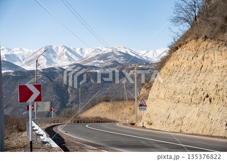 Winding mountain road with sharp turn signs cutting through a rocky hillside, with snow-covered peaks and deep valley in the background under blue sky Winding mountain road with sharp turn signs cutting through a rocky hillside, with snow-covered peaks and deep valley in the background under blue sky 135376822