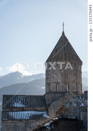 Stone drum, conical dome of the Cathedral of Saints Paul and Peter at Tatev Monastery rise above snow-dusted roofs with mountain ridges, winter, calm 135376945
