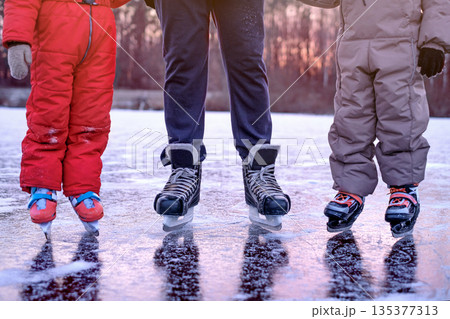Skating shoes of children and dads in close-up standing on the ice of a frozen lake at sunset at dusk 135377313